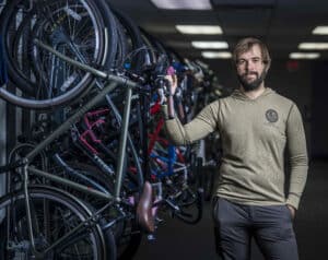 A man with a beard stands confidently next to a rack of colorful bicycles in a well-lit indoor space. He wears a beige sweater and dark pants.