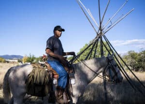 A man wearing a black cap and t-shirt rides a saddled horse past a set of wooden tipi poles in a grassy field under a clear blue sky.