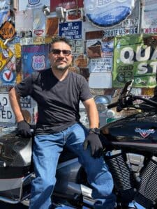 Man with sunglasses, black t-shirt, and jeans on a motorcycle by a wall covered in flyers.