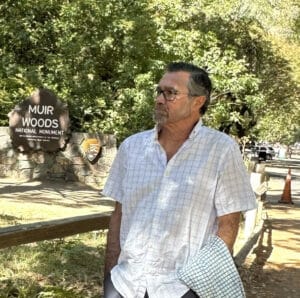 A man with glasses and a white, short-sleeve, button-down shirt is looking to the left. Behind him are leafy trees and a sign that reads Muir Woods National Monument.