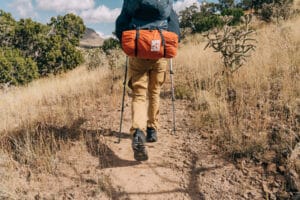 image of a man from the shoulders down and from behind, wearing black hiking shoes, tan hiking pants, black jacket, and carrying a large black pack on his back and a smaller orange pack beneath. The man is using metal hiking poles and is traversing a southern desert environment with weeds, pinon, and cholla cactus.