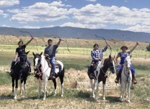 4 youth on horseback posing with archery bows held high with field, mountains, and clouds in the background.