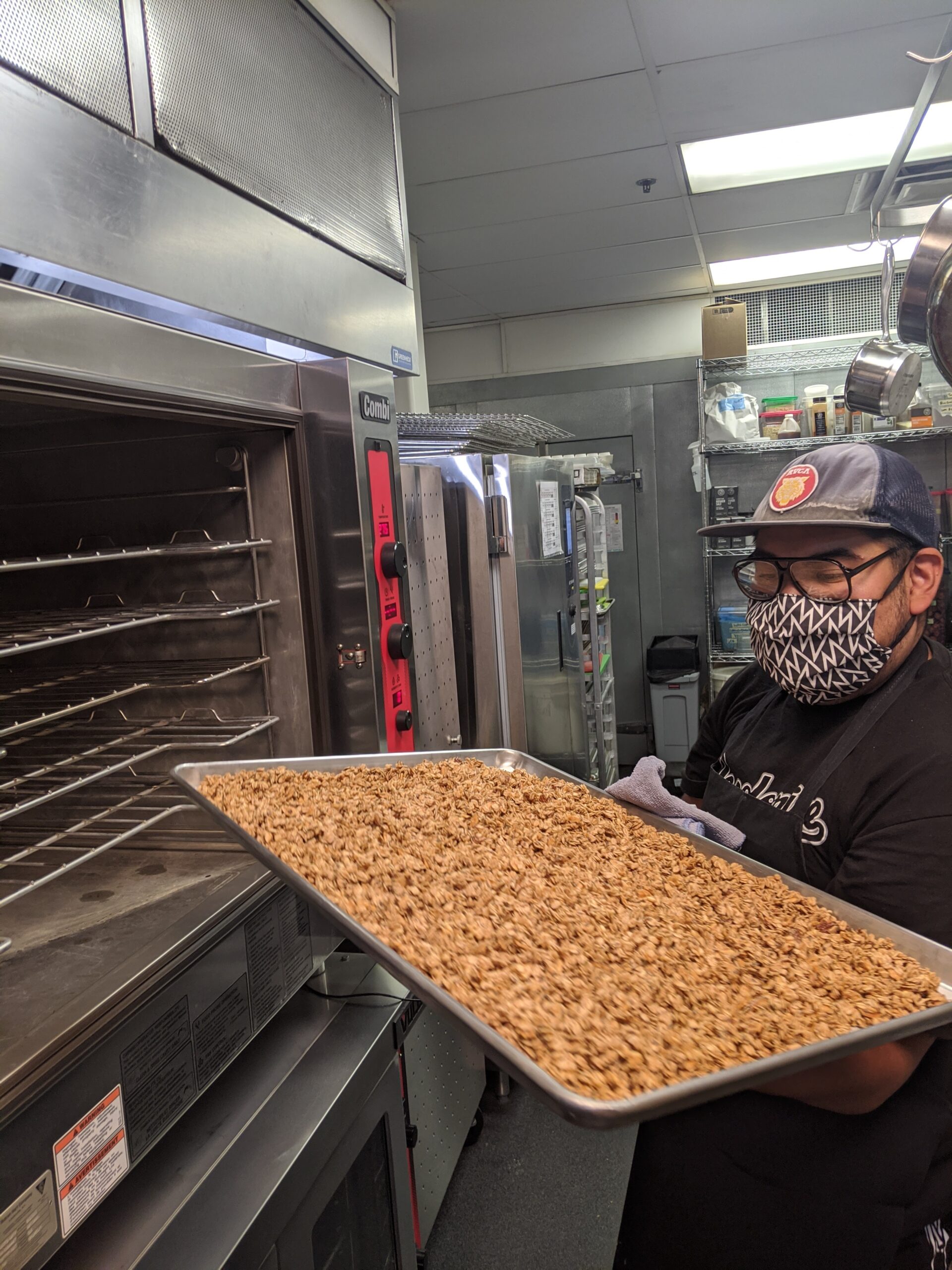 Kitchen worker stands with a full tray of fresh granola outside the commercial oven