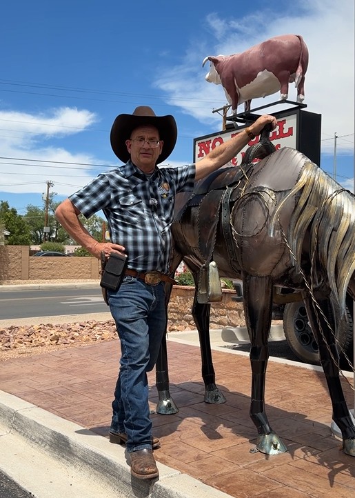 Brett Rizzy, Owner of No Bull Prime Meats, poses in a cowboy hat, plaid shirt, and jeans beside his metal structure of a horse.
