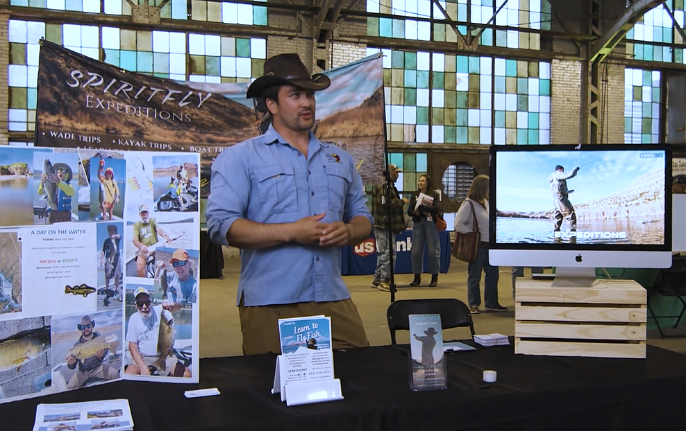 Man in blue shirt and black cowboy hat stands behind a vendor booth showcasing his business.