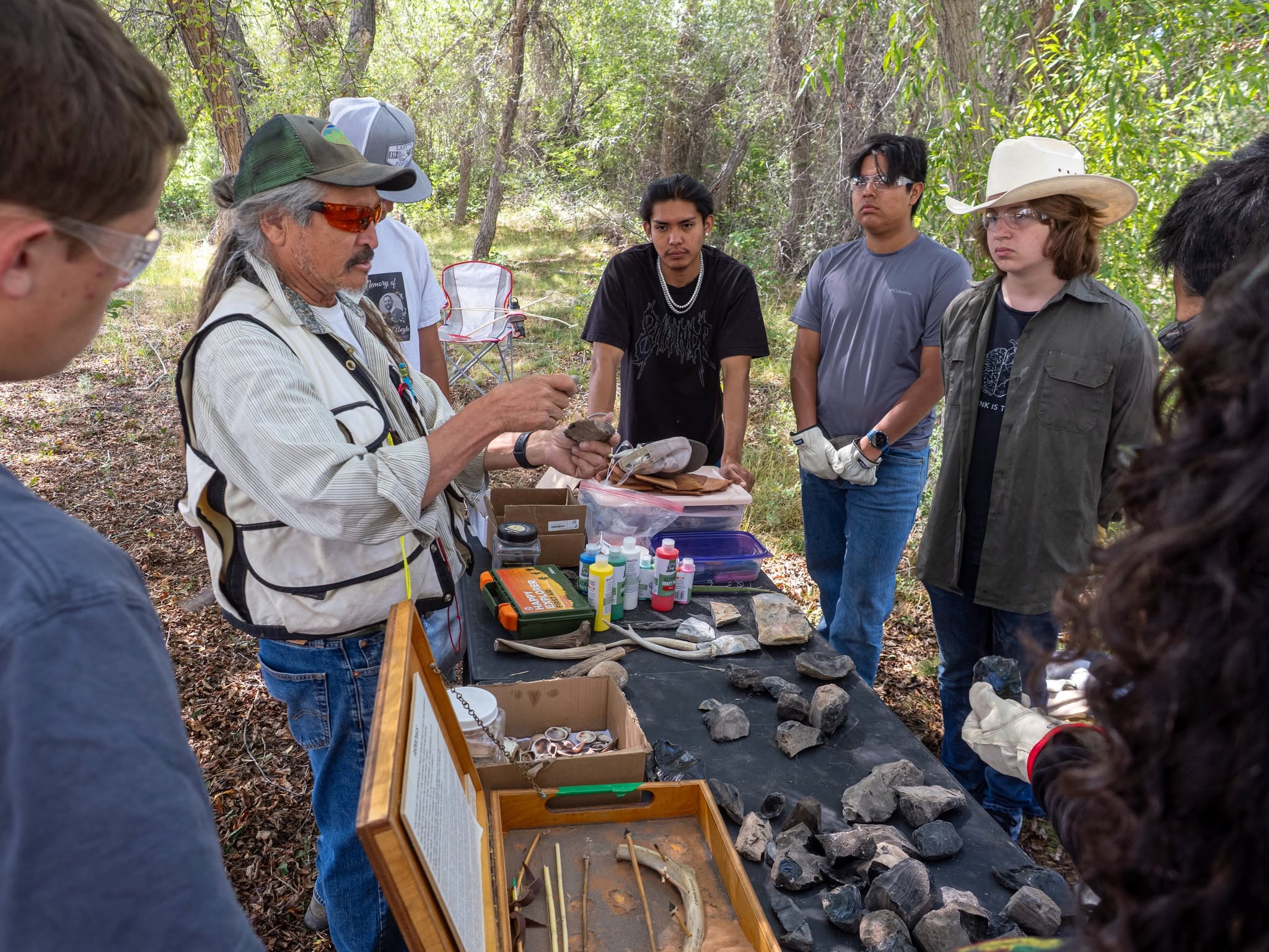 An educator in outdoor garb stands behind a table with rock displays surrounded by several teenagers.