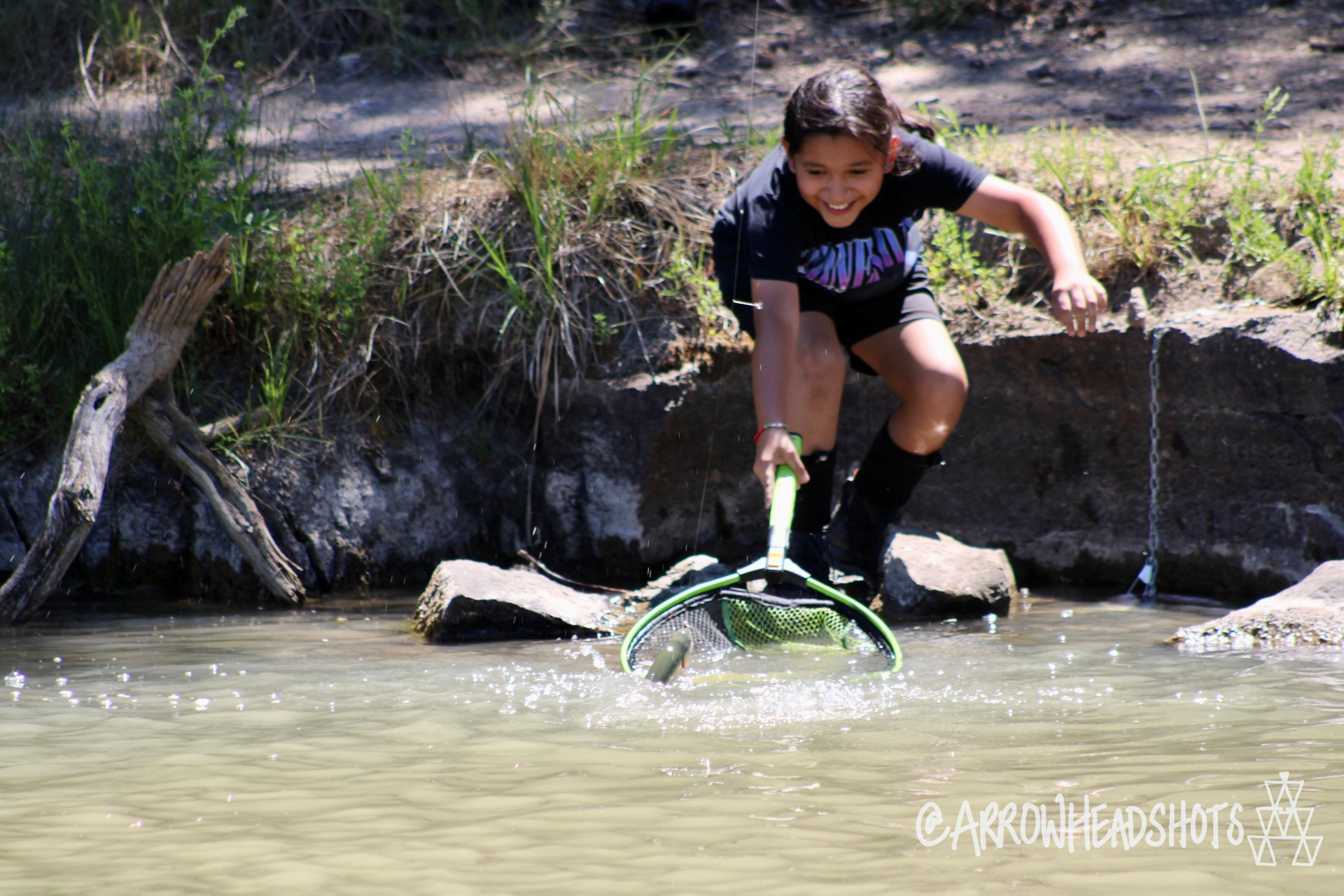 A young person is smiling by the side of a river while holding a net in the water.
